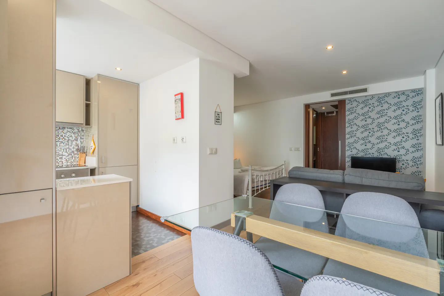 A bright apartment interior with a kitchen, dining area, and living room. Gray chairs surround a glass table. A gray sofa faces a TV with patterned wallpaper.