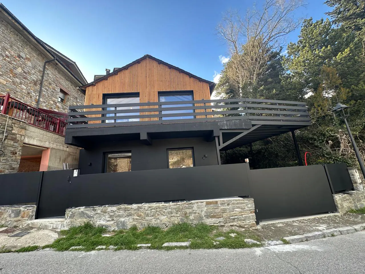 Modern two-story house with a wood-paneled top floor, black exterior, and a large balcony, set against a backdrop of trees and blue sky.