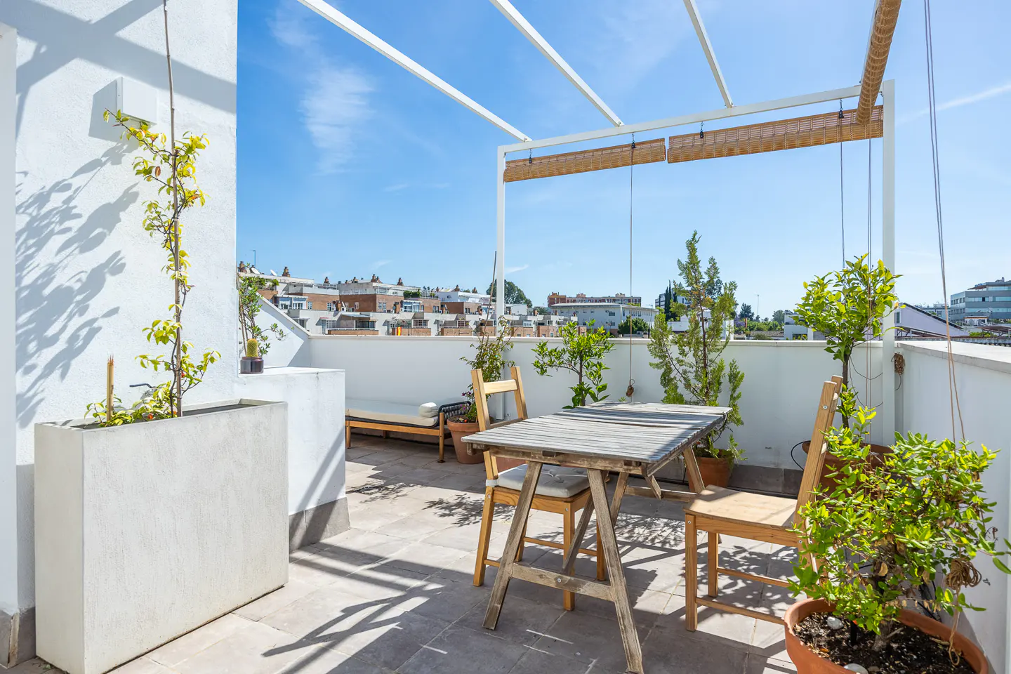 Rooftop patio with a wooden table, chairs, and plants in pots. White walls and a pergola frame the blue sky and city view.
