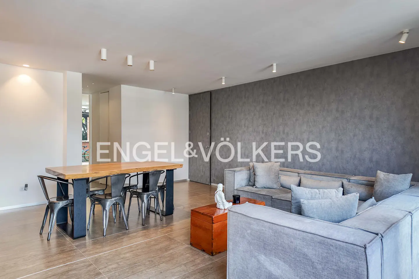 Modern living room with a wooden dining table, metal chairs, a large grey sectional sofa, and a textured grey accent wall.