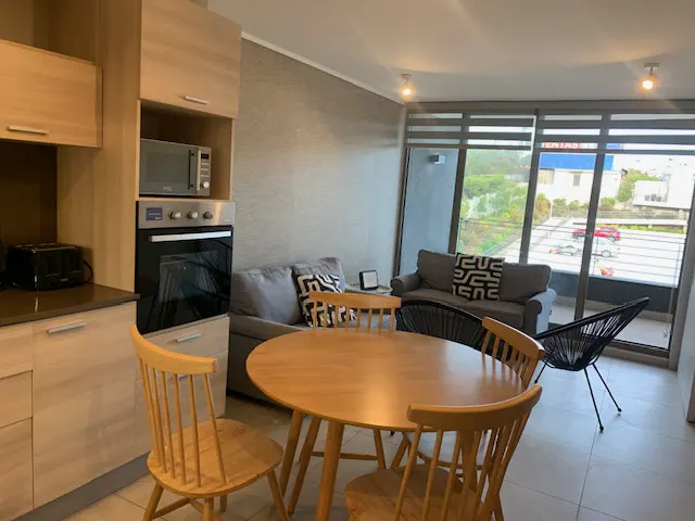 A bright apartment interior with a round wooden table and chairs in the foreground, a gray sofa, and a kitchen area with appliances. Large windows offer a view of the outside.