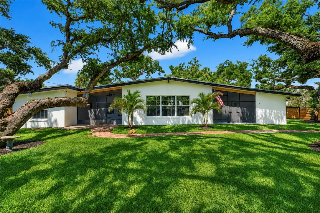 A white, one-story house with gray doors and trim, framed by large trees and a green lawn under a blue sky.