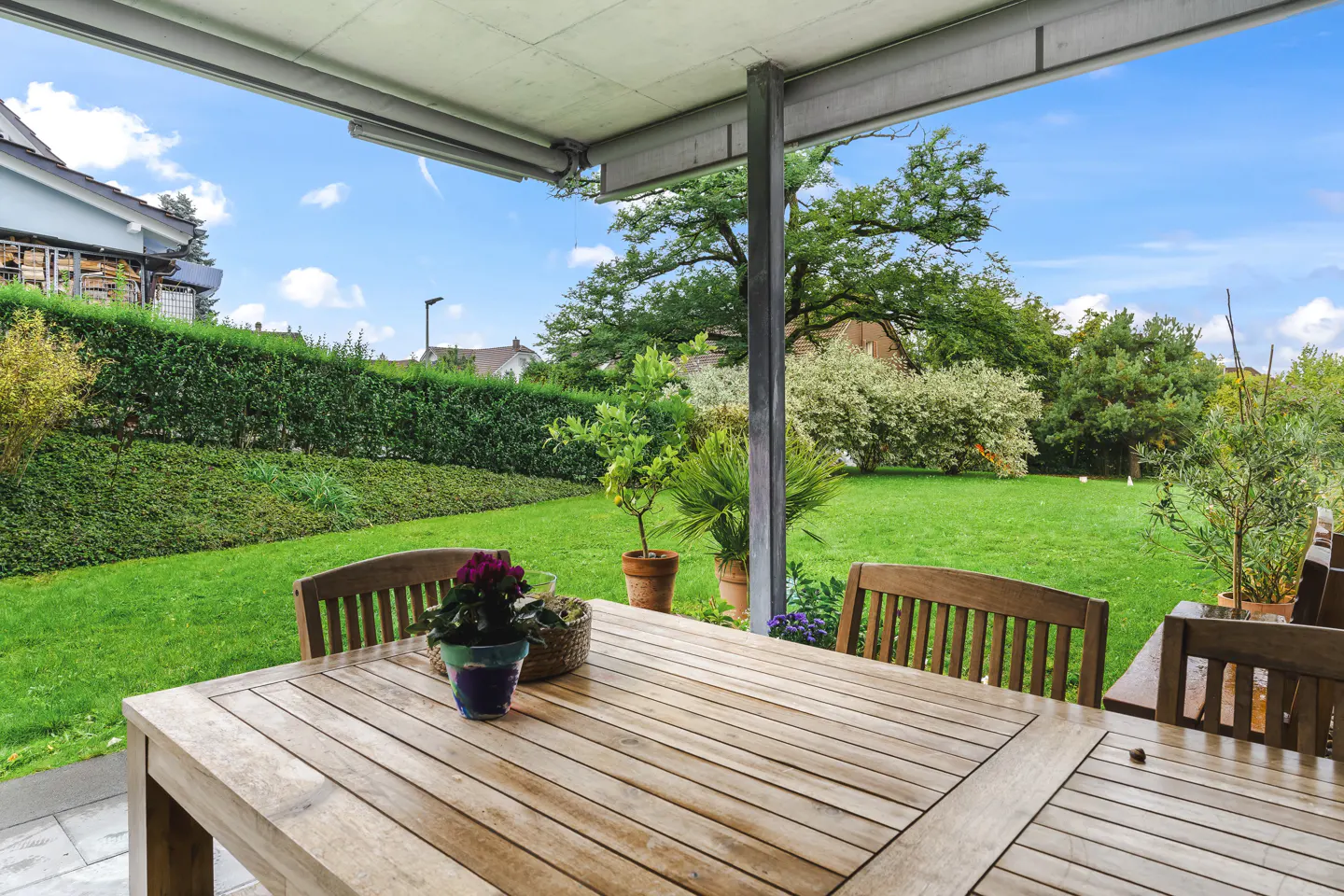 Covered patio with a wooden table and chairs overlooking a green lawn, trees, and blue sky.