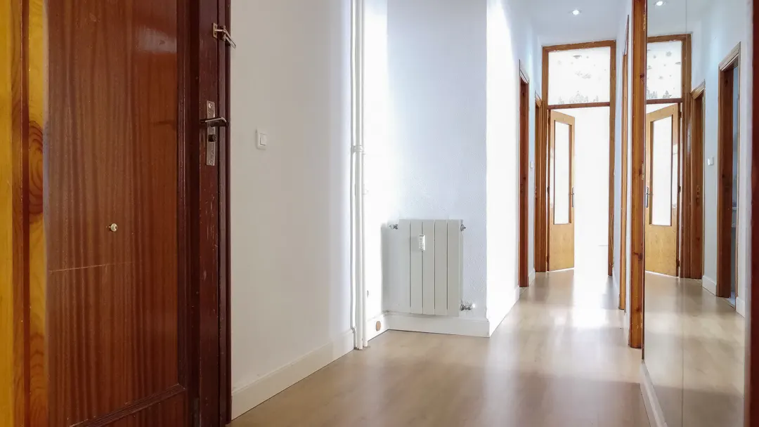 A bright hallway with wood floors, white walls, and brown doors. A radiator is on the wall.