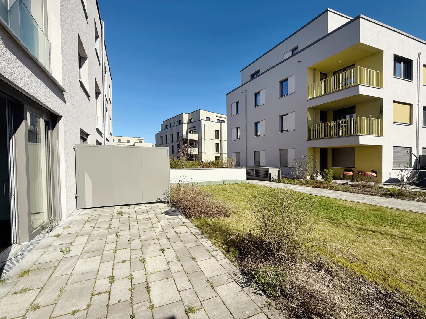 Apartment complex exterior with gray patio, green lawn, and modern white buildings under a clear blue sky.