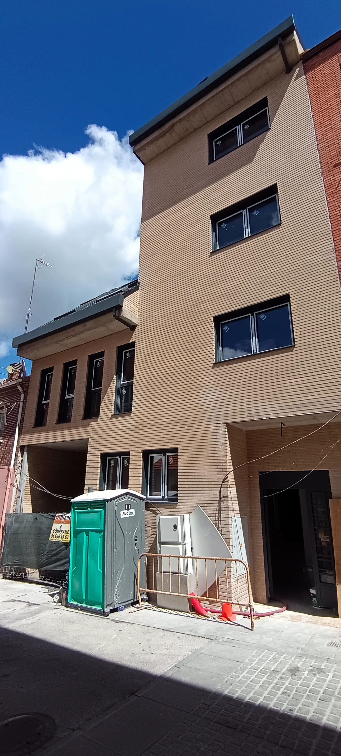Exterior view of a three-story tan brick building with black-framed windows under a blue sky with white clouds. A green portable toilet sits on the street.