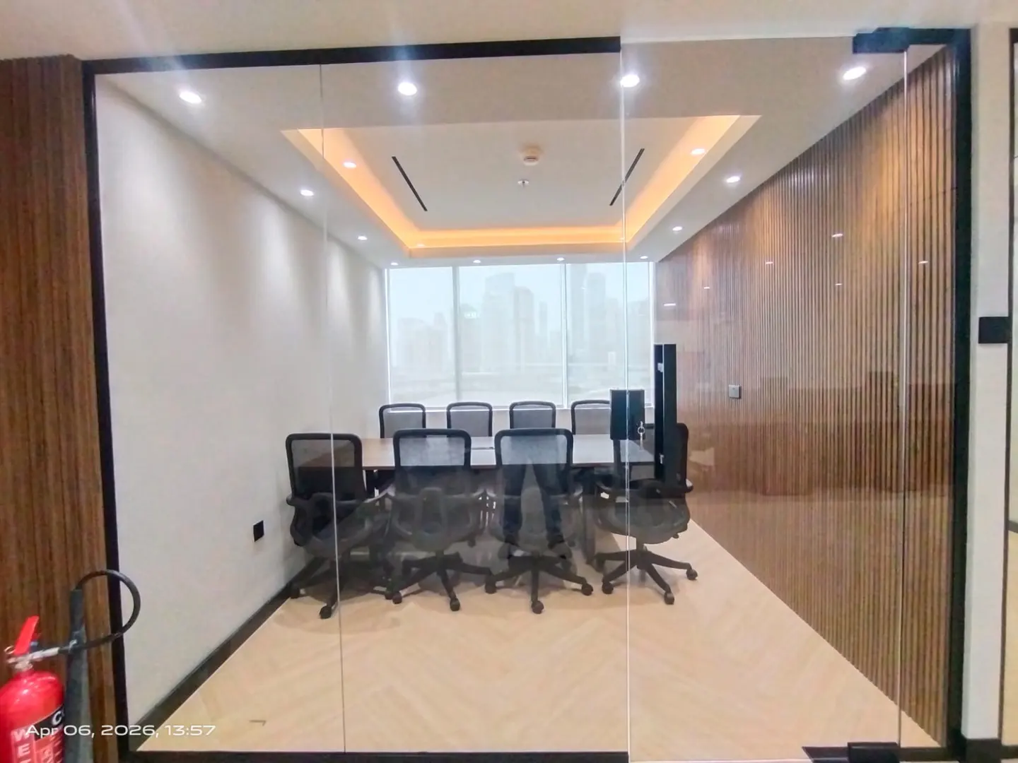 A modern conference room with a long table, black chairs, and a city view through a large window. The room has wood paneling and a glass door.