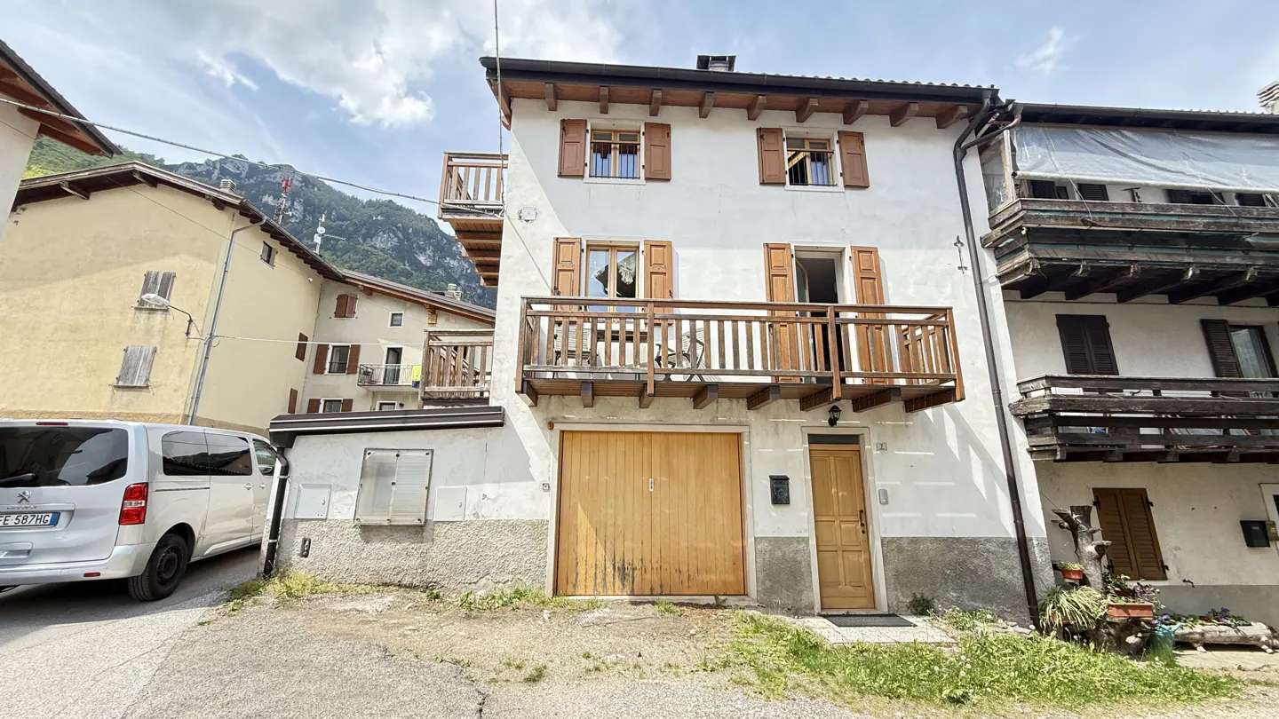 Exterior of a white two-story house with brown shutters and balconies, set in a mountain village. A silver van is parked nearby.
