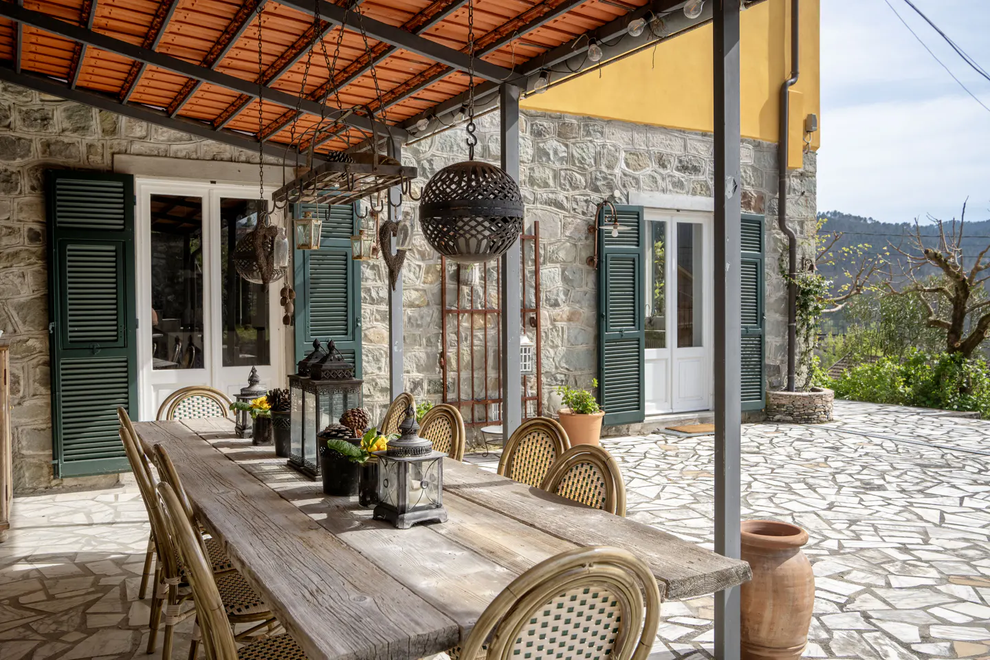 Outdoor patio with a long wooden table, wicker chairs, lanterns, and stone flooring. The building has stone walls and green shutters.