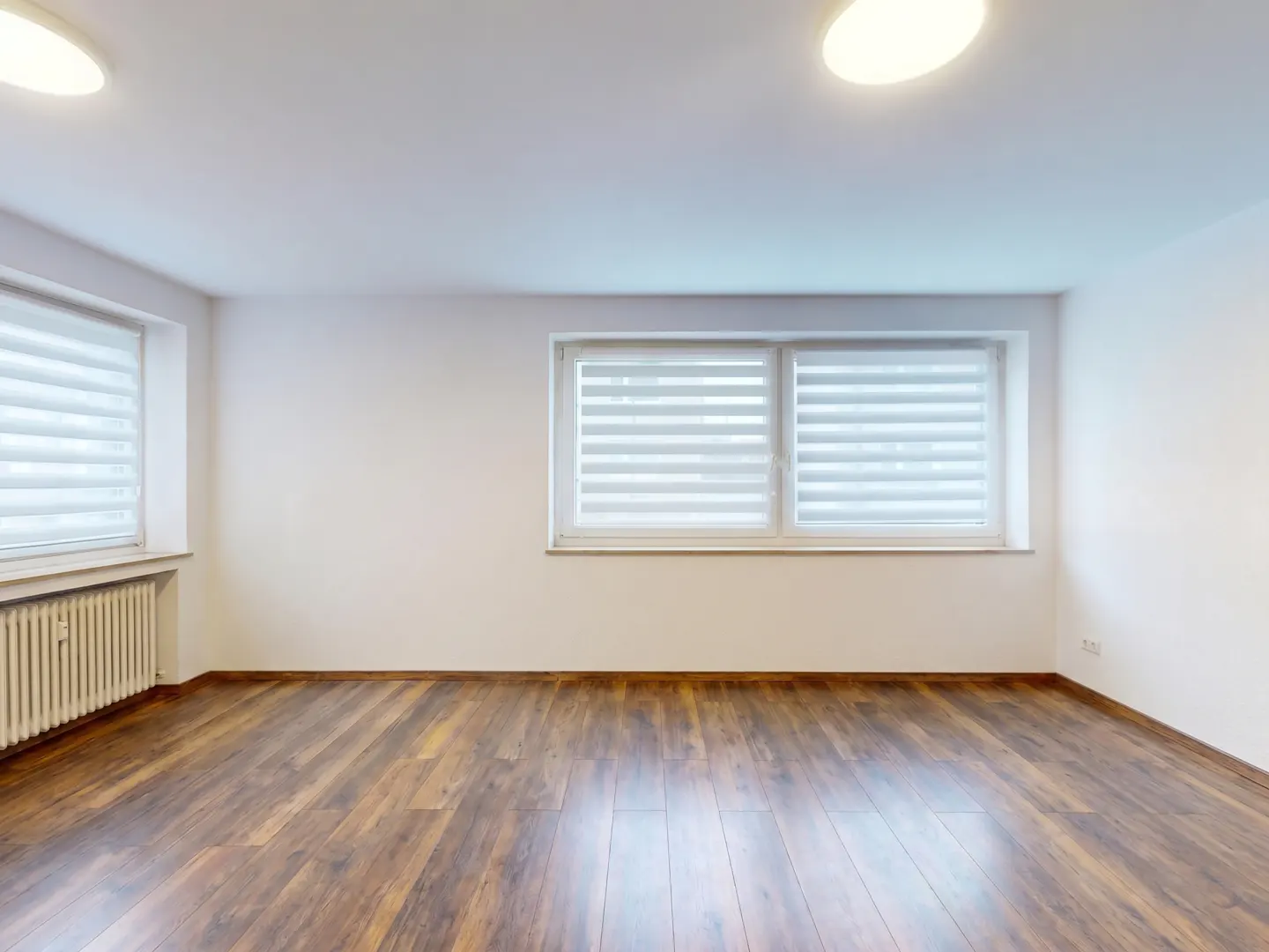 Bright, empty room with wood floors, white walls, and two windows with white blinds. Two ceiling lights illuminate the space.