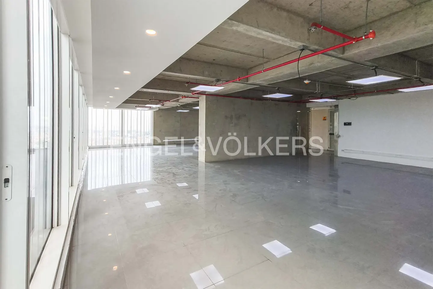 Bright, empty office space with concrete ceiling, red pipes, and large windows. Gray floors reflect light. Engel & Völkers logo visible.