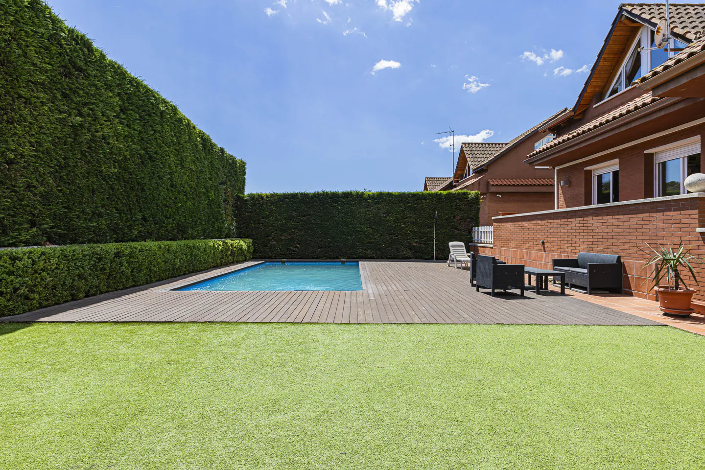 Backyard with a pool, wood deck, and green lawn. Tall green hedges line the back, with a brick house on the right. Blue sky above.