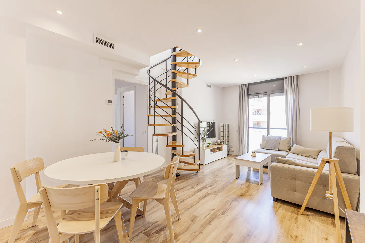 Bright living room with wood floors, white walls, and a spiral staircase. A white table with chairs sits near a beige sofa and a window with curtains.