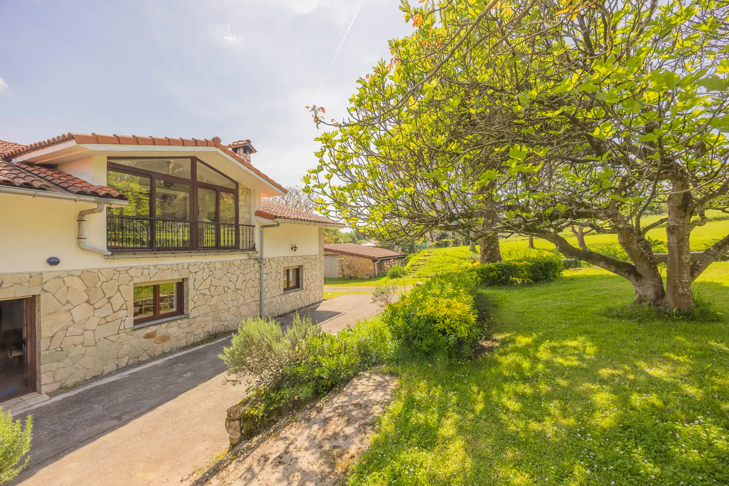 Exterior view of a stone house with a red tile roof, a balcony, and a large tree in the yard.
