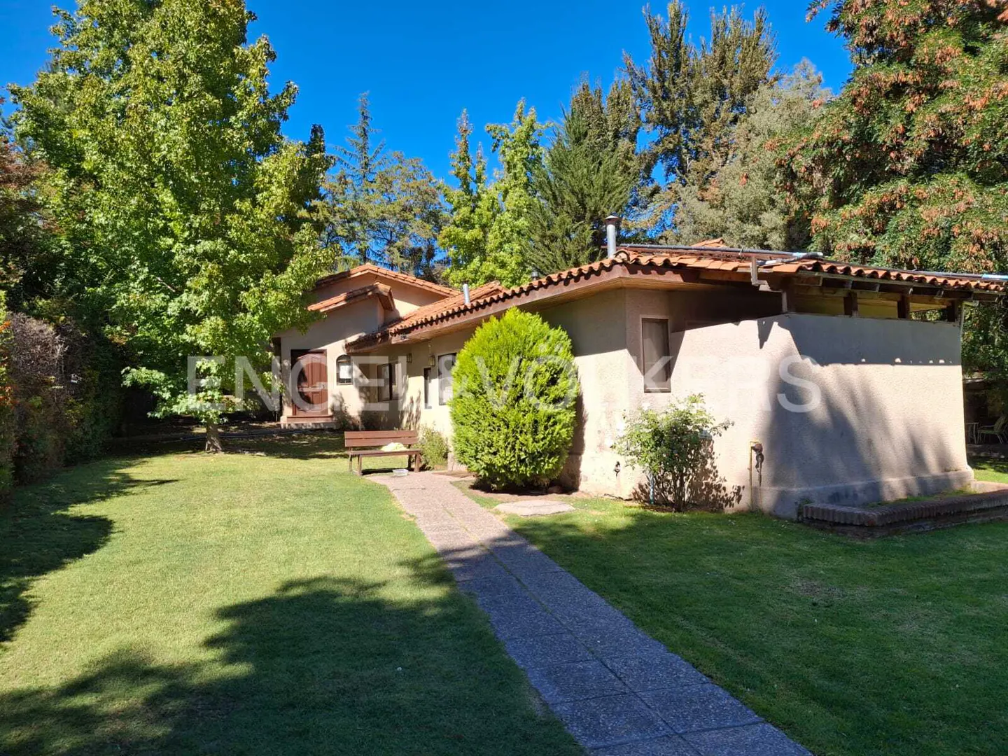 Exterior view of a beige house with a red tile roof, green lawn, trees, and a stone walkway under a blue sky.