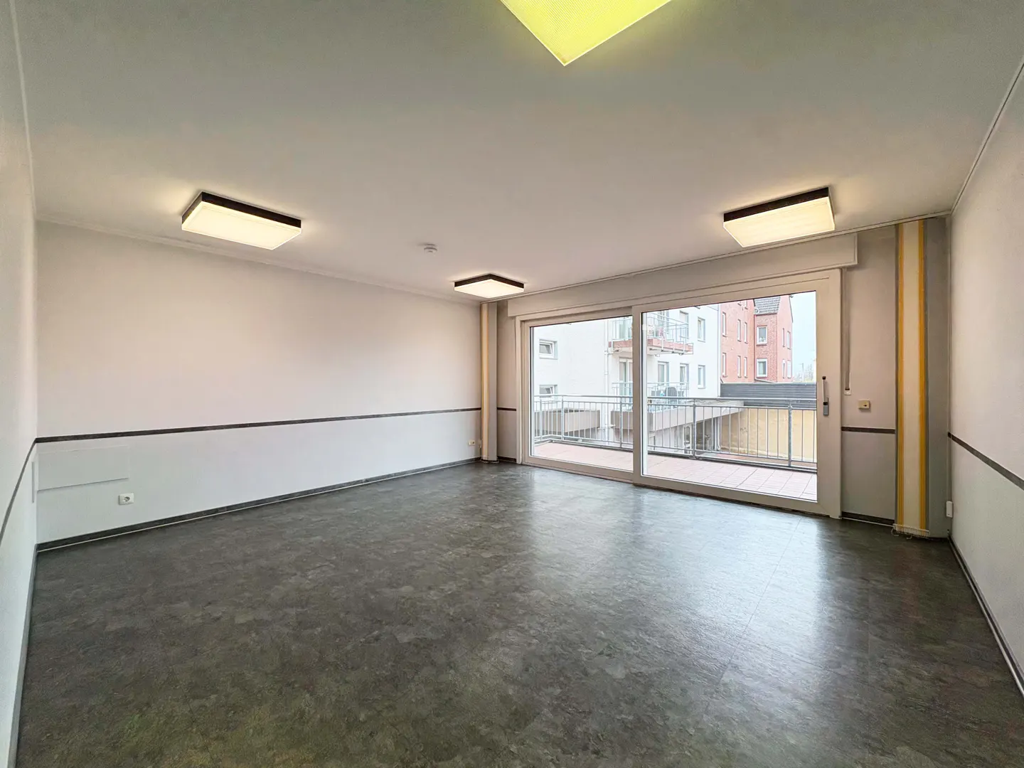 Empty room with gray floor, white walls with black trim, and sliding glass doors to a balcony. Three square ceiling lights illuminate the space.