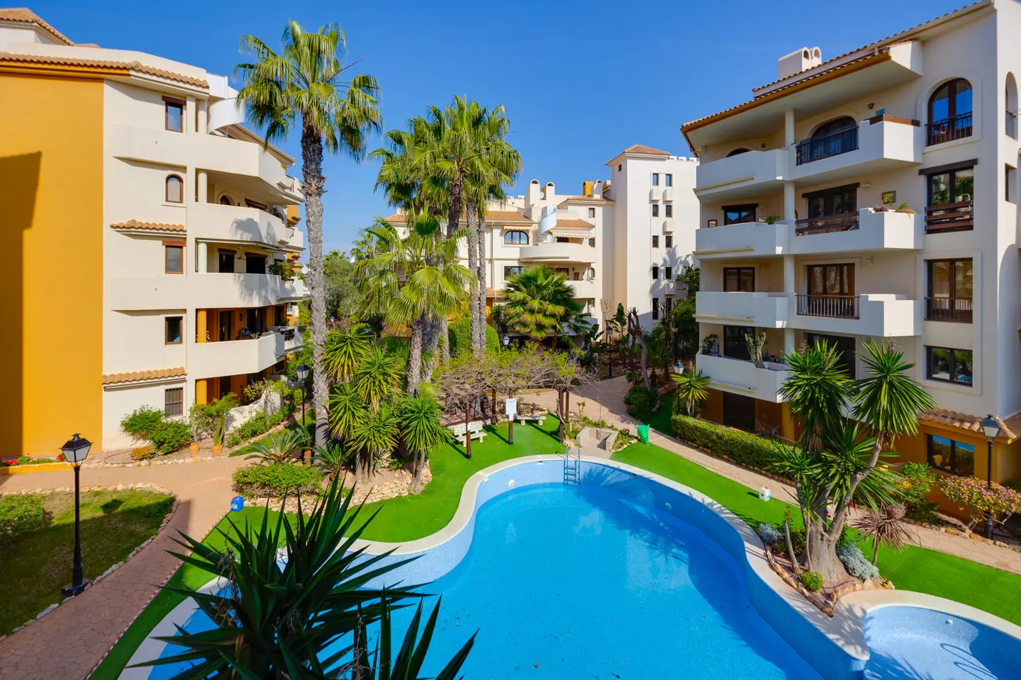Exterior view of a resort with a blue swimming pool, palm trees, and multi-story buildings under a clear blue sky.