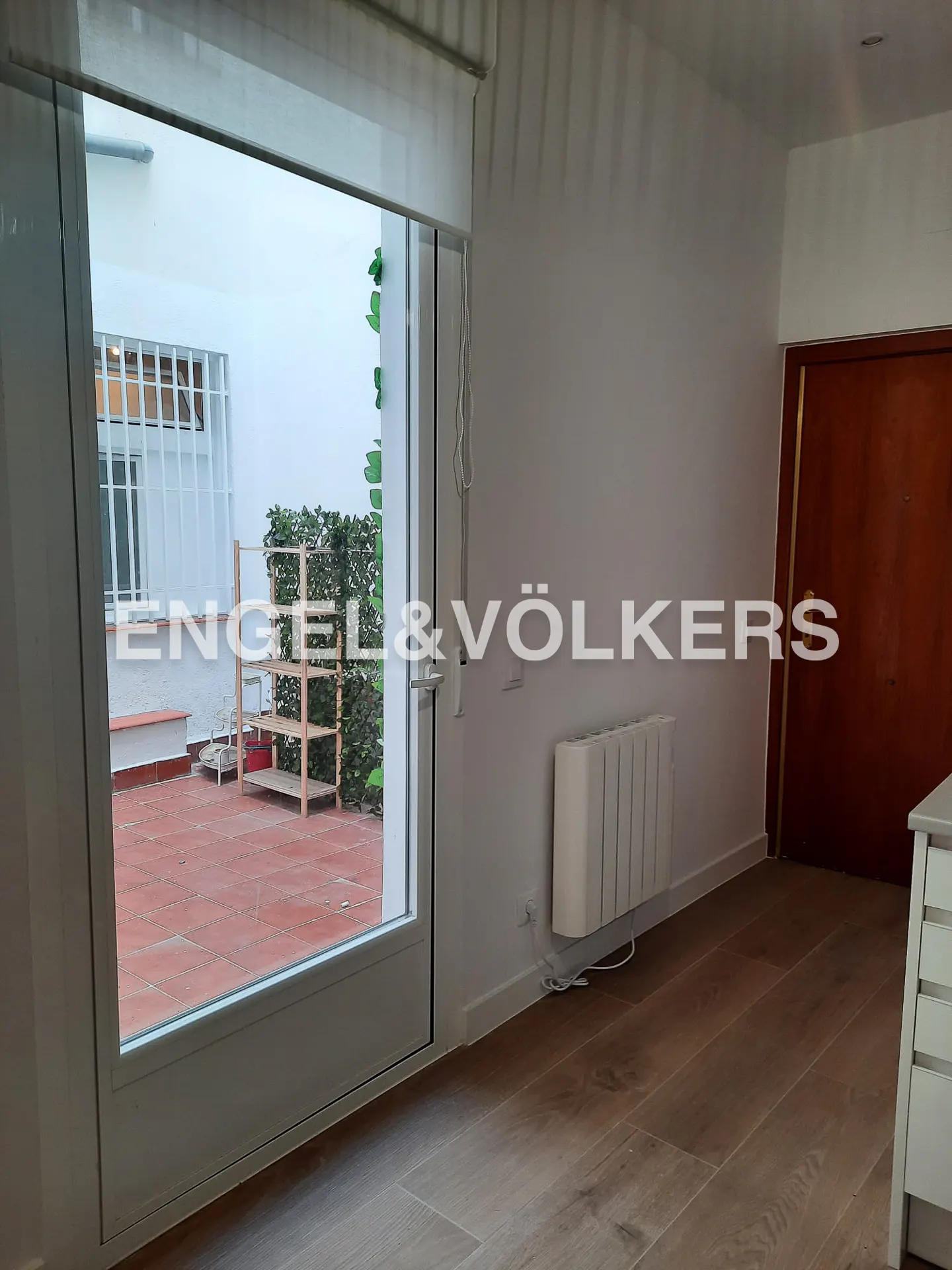 A bright room with wood floors, a white radiator, and a glass door leading to a red-tiled patio with greenery.