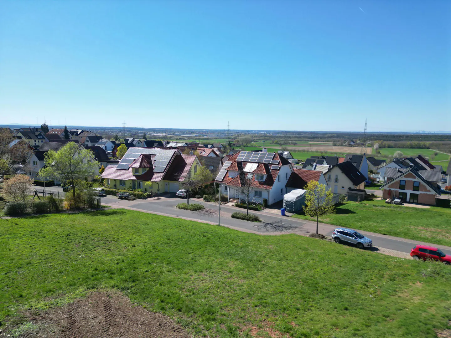 Aerial view of a suburban neighborhood with houses, green lawns, and cars under a clear blue sky. Solar panels are visible on some roofs.