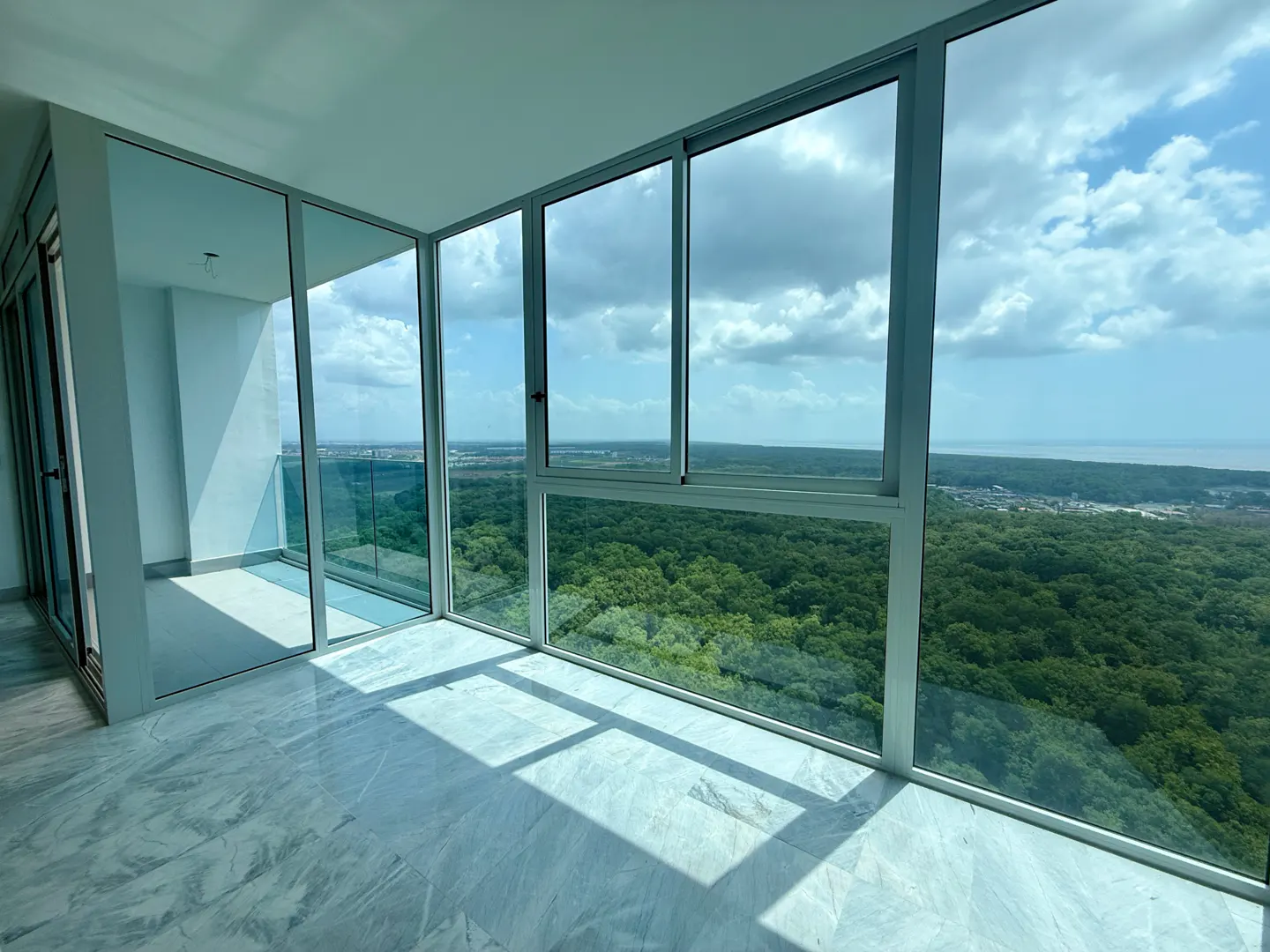 Bright room with marble floors and floor-to-ceiling windows overlooking a green forest and cloudy blue sky.