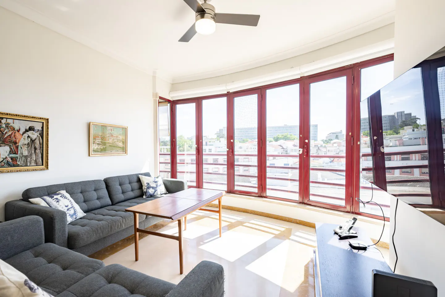 Bright living room with a grey sectional sofa, wooden coffee table, ceiling fan, and large windows with a city view.