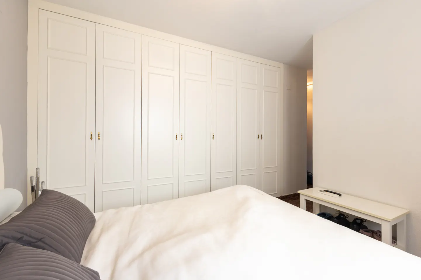 Bedroom with a white bed, gray pillows, and a wall of white closets with gold handles.
