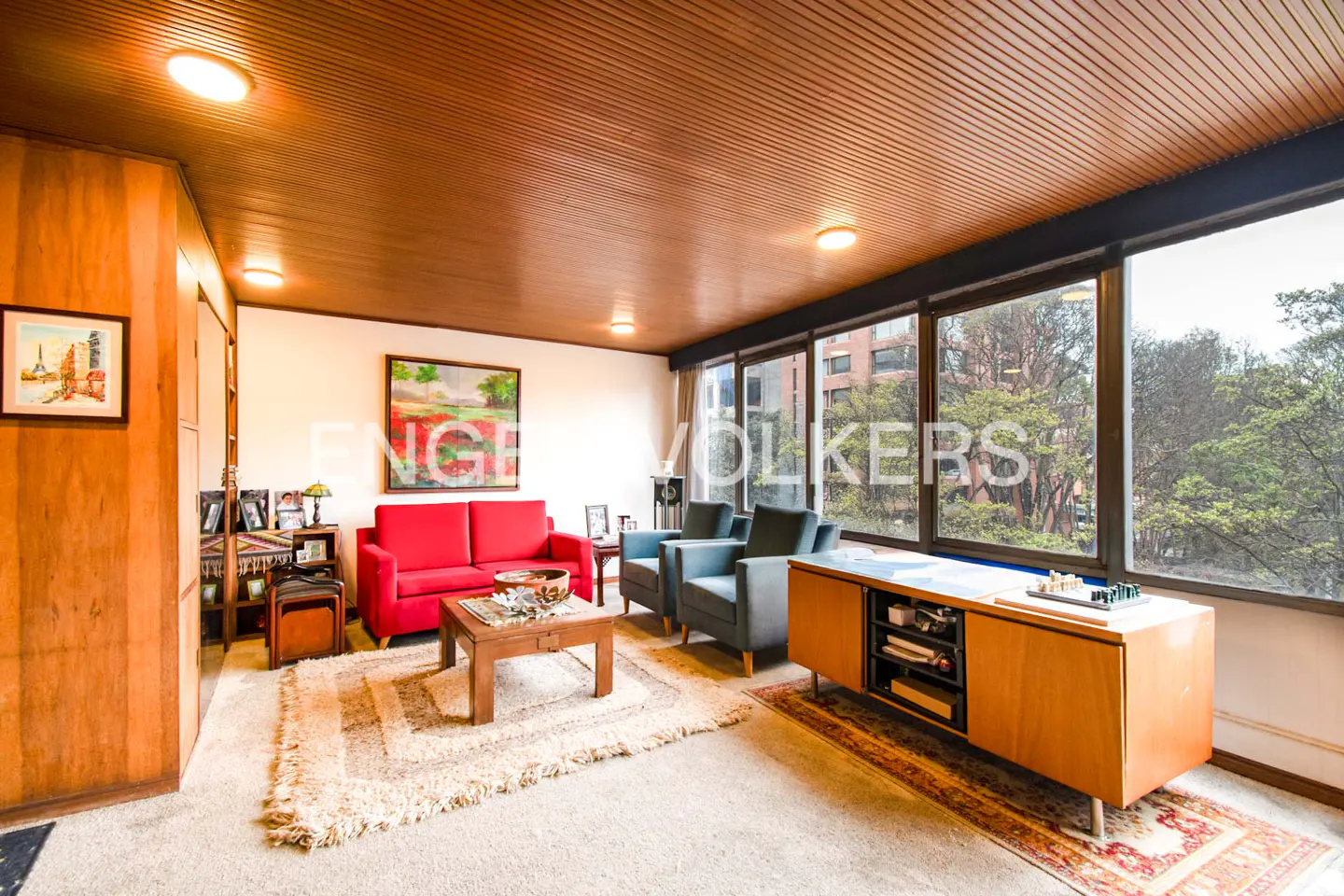 Mid-century living room with wood-paneled ceiling, red sofa, two grey armchairs, coffee table, and large windows.