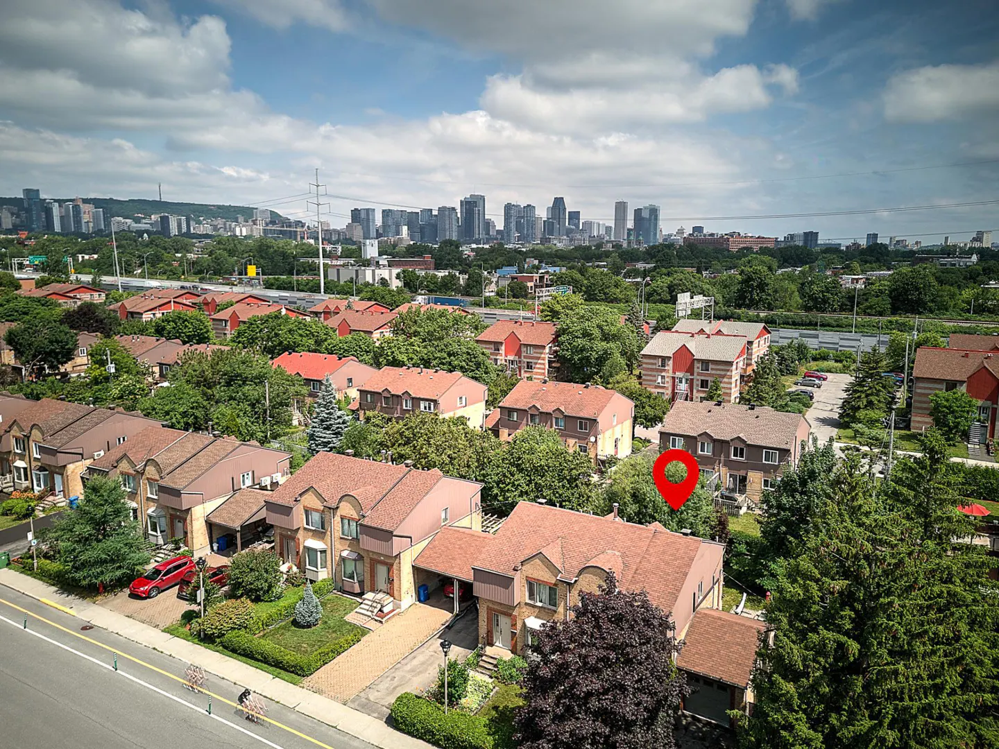 Aerial view of a suburban neighborhood with brown-roofed houses, green trees, and a red location pin. A city skyline is visible in the background under a cloudy sky.