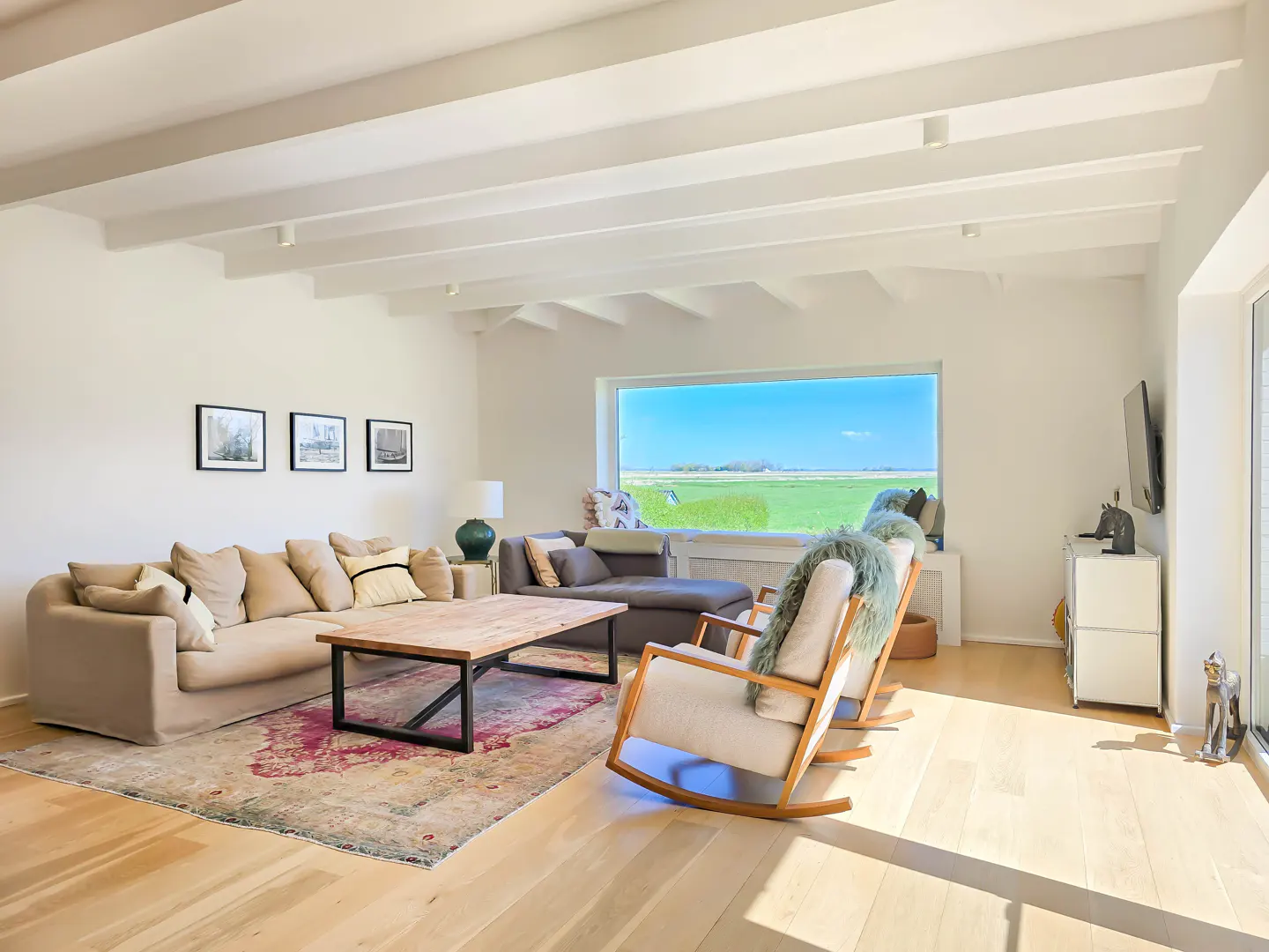 Bright living room with white walls, wood floors, and exposed beams. A large window overlooks a green field. Beige sofa, rocking chairs, and a wood coffee table furnish the space.