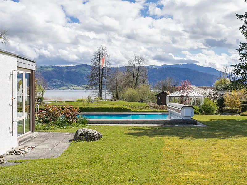 A backyard with a pool, green lawn, and lake view. Mountains are visible in the background under a cloudy sky.