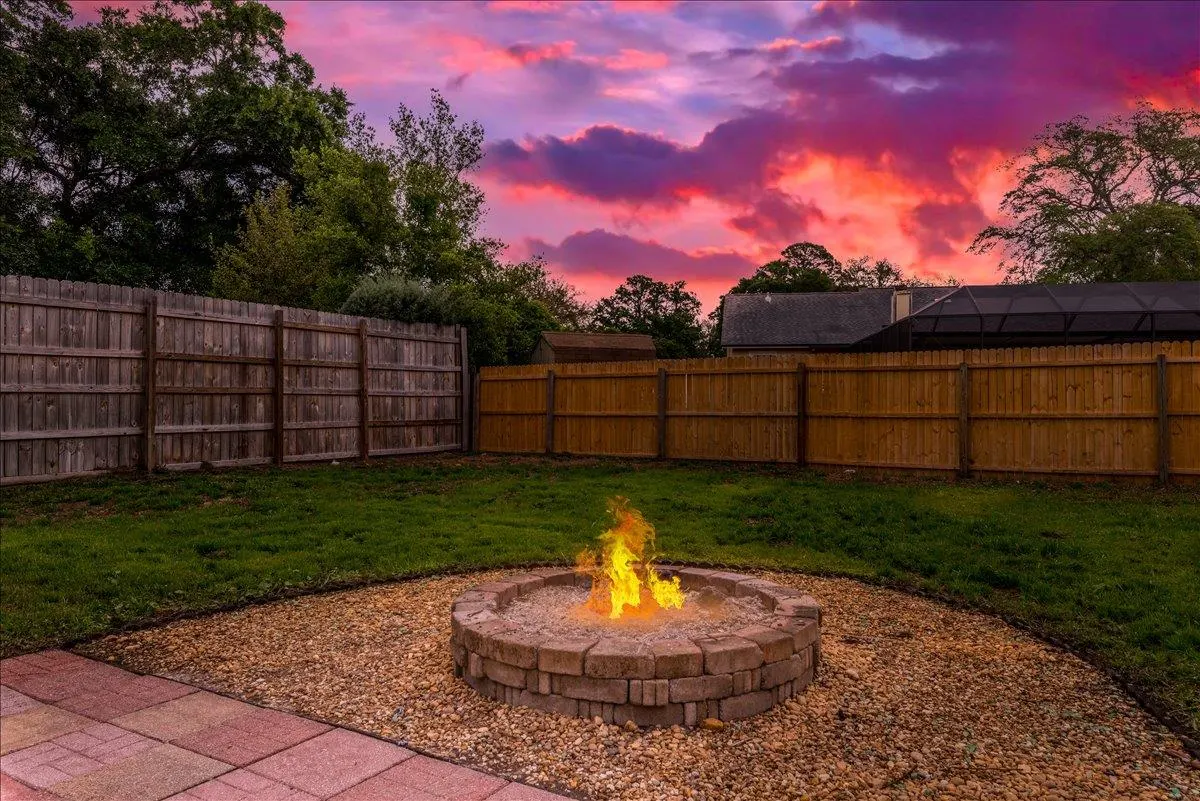 Backyard with a stone fire pit ablaze, surrounded by gravel and grass. A wooden fence encloses the yard under a vibrant pink and orange sunset.