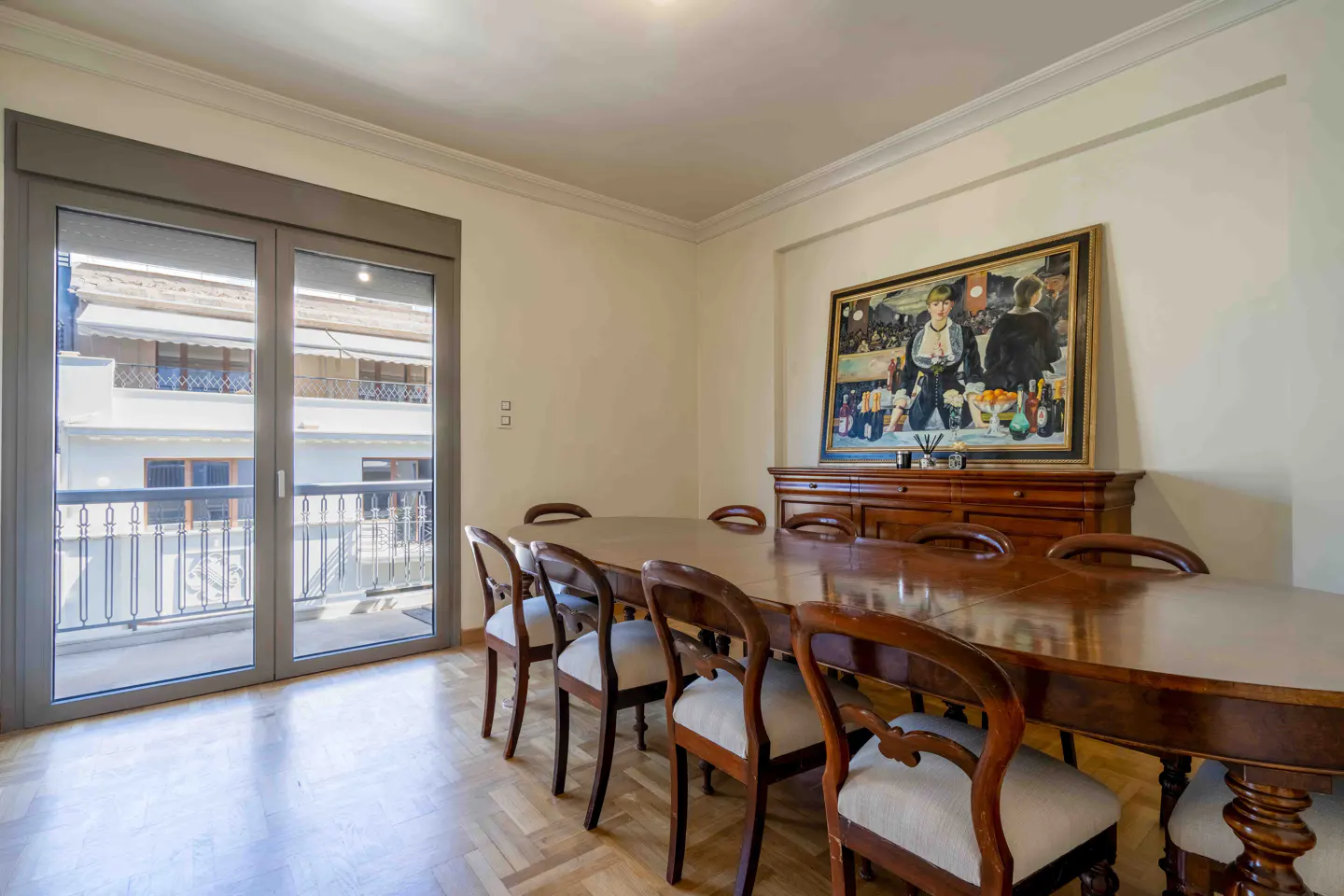 Dining room with a long wooden table, chairs, and a painting above a wooden sideboard. A glass door leads to a balcony with a view of buildings.