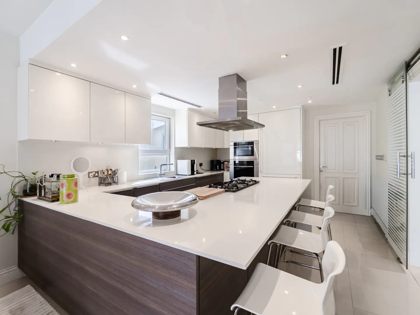 Bright, modern kitchen with white cabinets, a large island with a gas cooktop, and white bar stools. Stainless steel appliances and a range hood are visible.