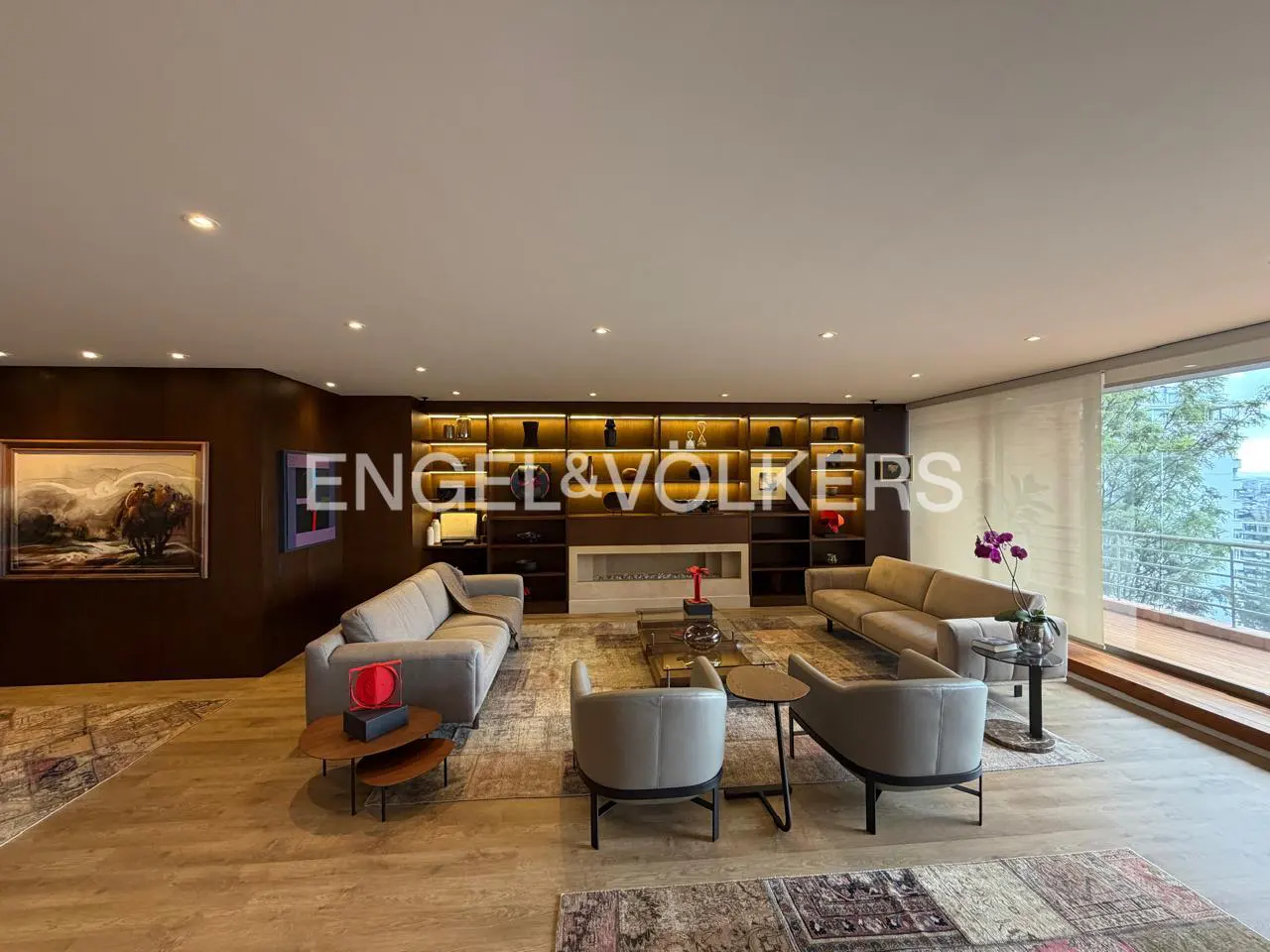 A spacious living room with light wood floors, gray sofas, armchairs, and a large built-in bookshelf with a fireplace.