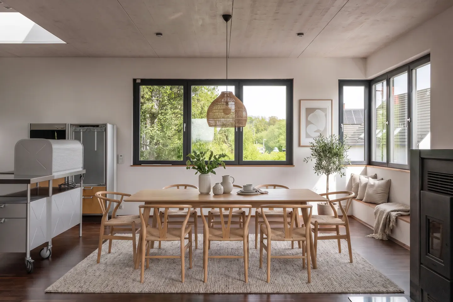 Bright dining room with a light wood table, woven chairs, and a woven pendant light. Large windows offer a view of green trees.