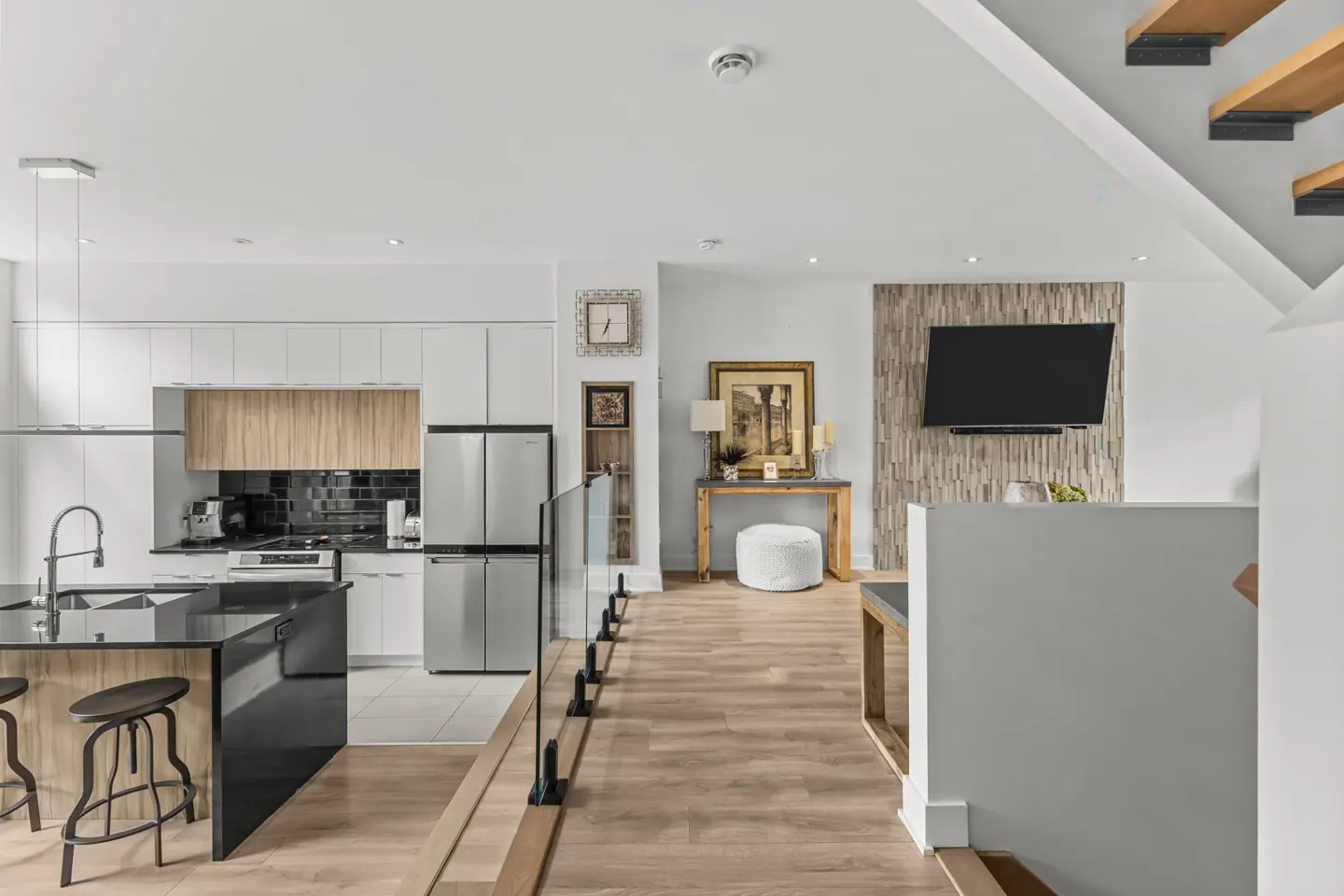 A modern home interior with a kitchen island, stainless steel appliances, and light wood floors. A TV is mounted on a textured wall.