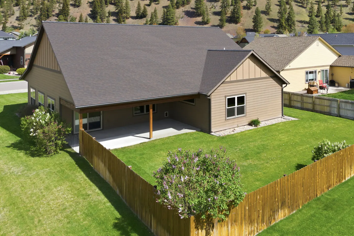 Aerial view of a tan house with a brown roof, a covered patio, and a green lawn enclosed by a wooden fence.