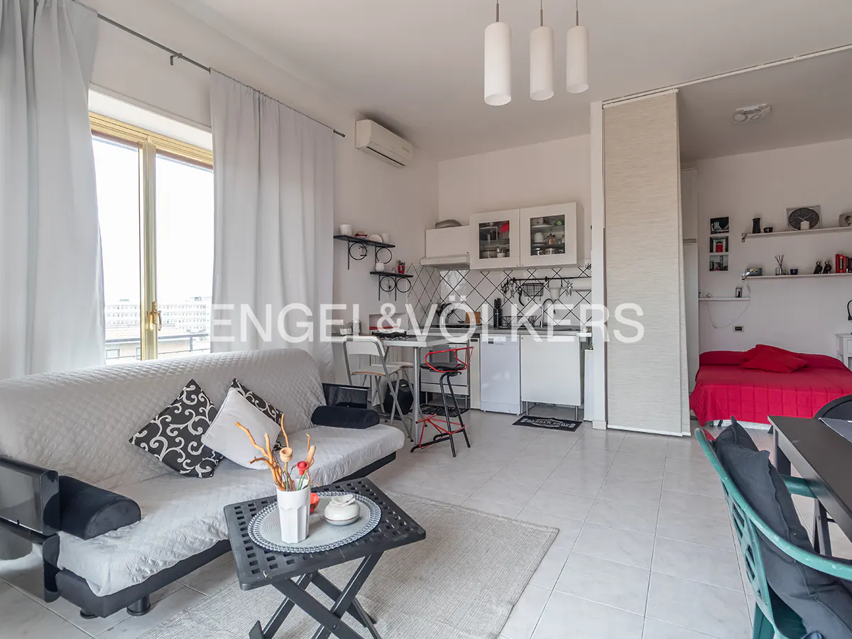 Bright studio apartment with white walls, tile floors, and a sofa. A small kitchen and a red bed are visible in the background.