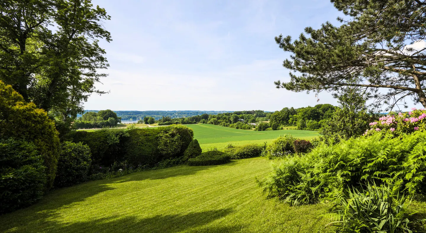 A lush green lawn slopes down to a view of fields and trees under a blue sky.