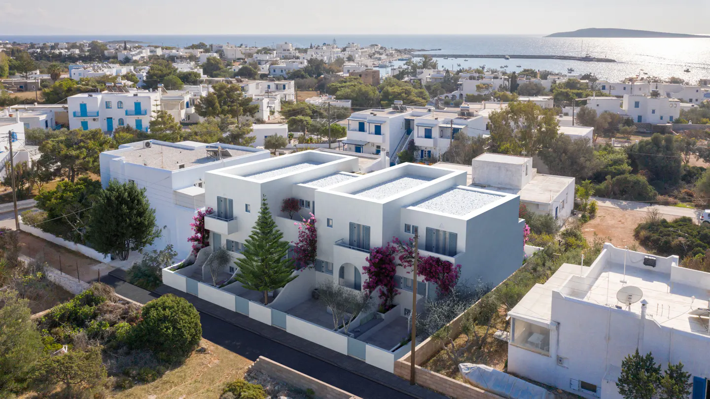 Aerial view of white villas with blue accents, surrounded by lush greenery and the sea in the background.