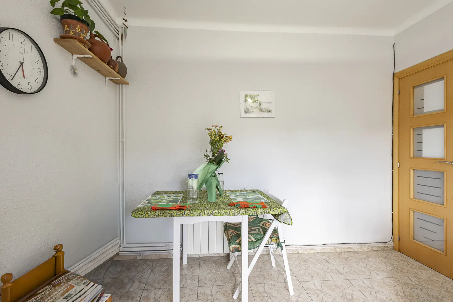 A bright room with a white table set for two, featuring a green patterned tablecloth and a vase of flowers. A wooden door is on the right.