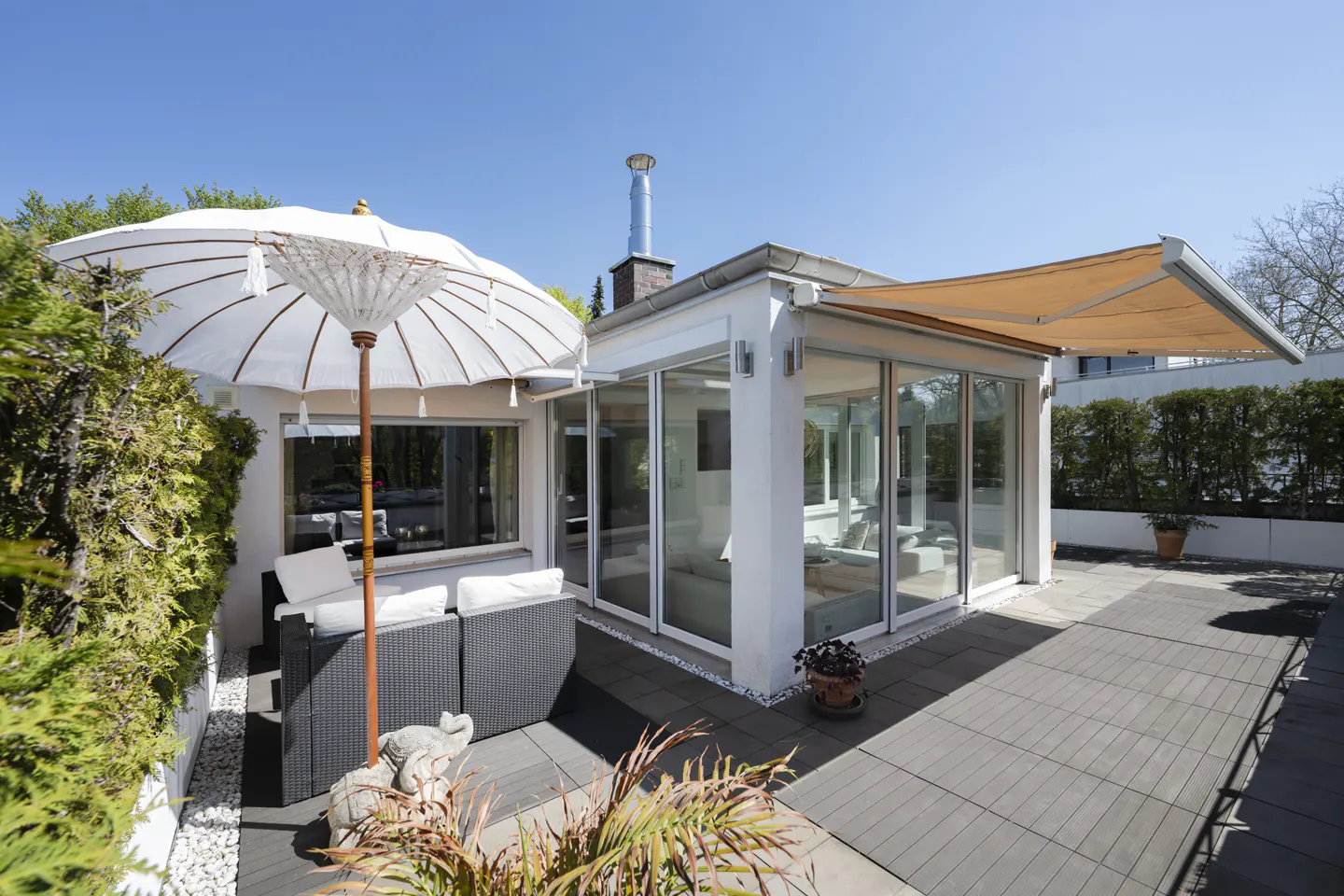 Modern patio with a white umbrella, wicker sofa, and glass-walled sunroom under a clear blue sky.