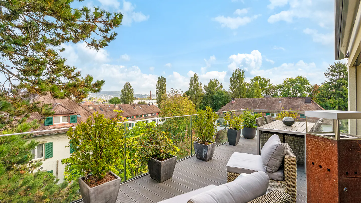 Balcony view with outdoor seating, potted plants, and a glass railing overlooking houses and trees under a blue sky.