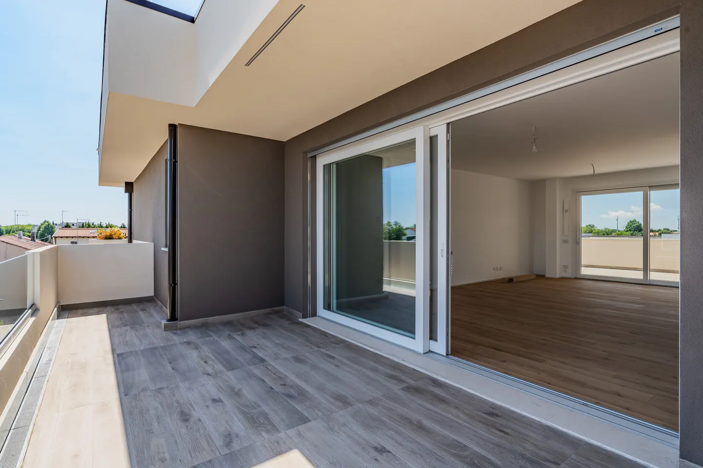 A modern apartment balcony with gray wood-look tile, sliding glass doors, and a view of the neighborhood.