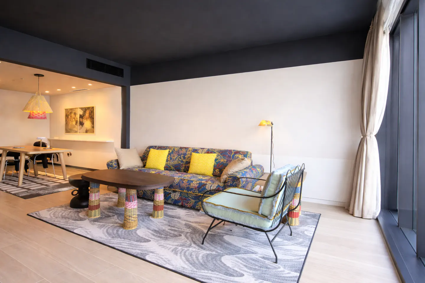 Living room with floral sofa, yellow pillows, and a light blue chair on a gray wave-patterned rug. A dining area is visible in the background.