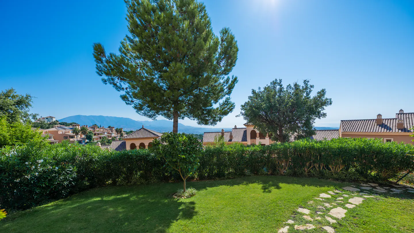 Lush green lawn with stone path, manicured hedges, and trees under a clear blue sky. Houses and mountains are visible in the background.