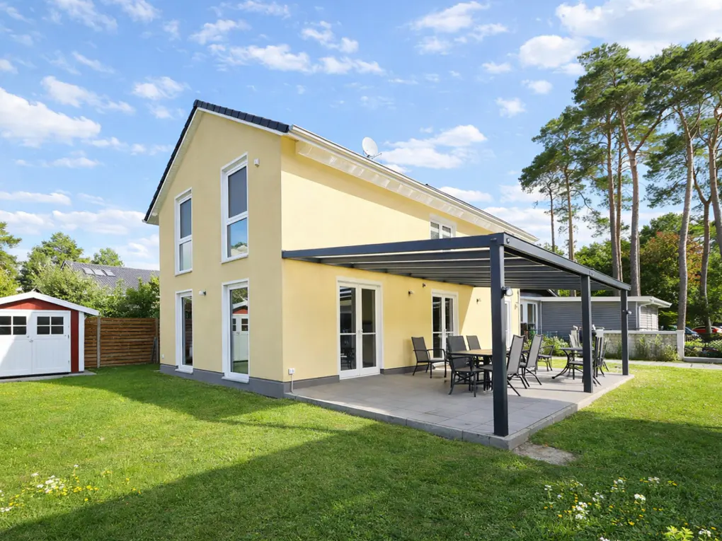 Two-story yellow house with a black pergola covering a patio with a table and chairs. A red and white shed sits on the green lawn.