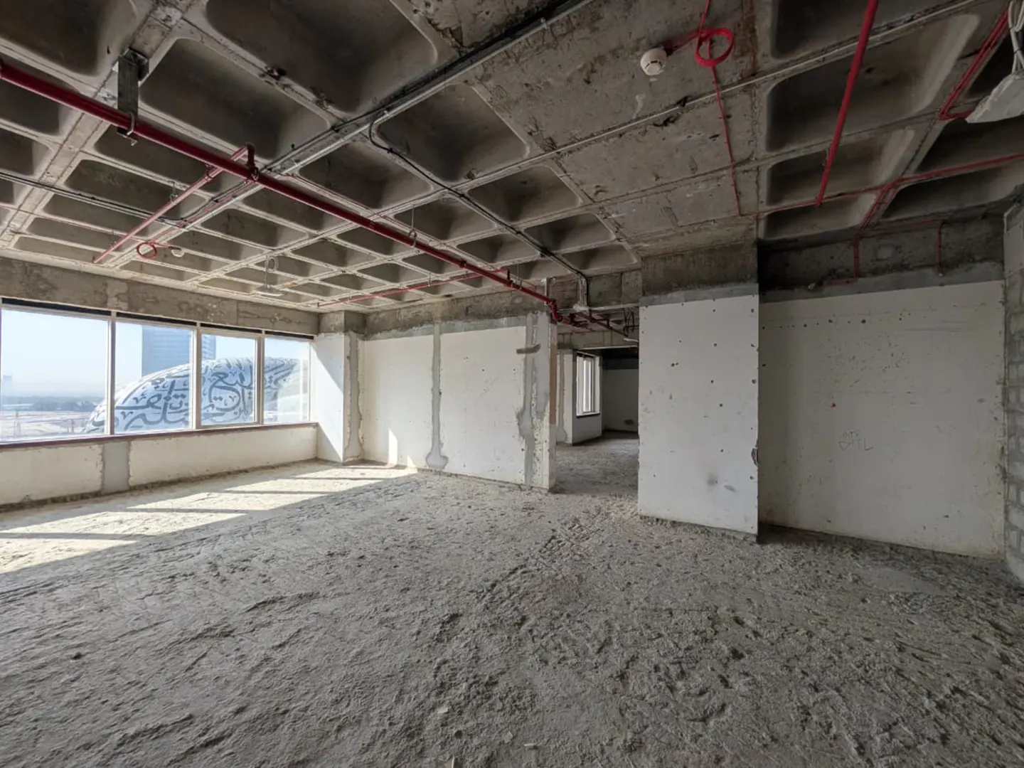 Unfinished office space with concrete floors, walls, and ceiling. Red pipes run across the ceiling. Windows show a building in the distance.