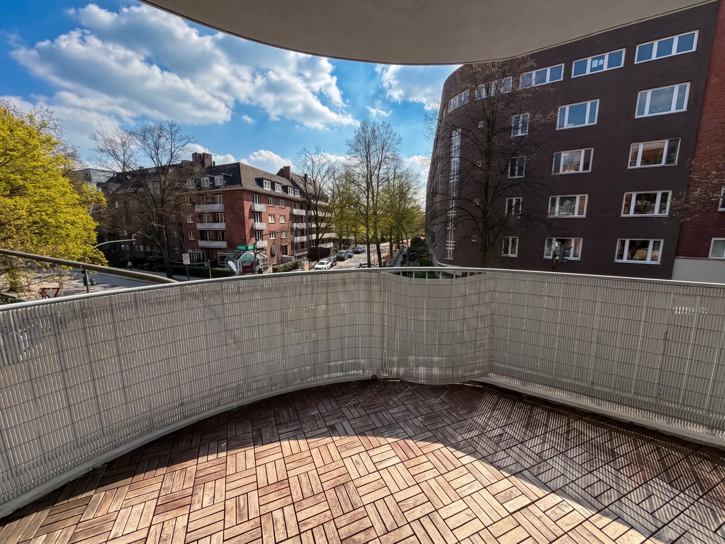 Balcony view with wood-look tile flooring and a gray privacy screen. Buildings and trees are visible in the background under a blue sky.