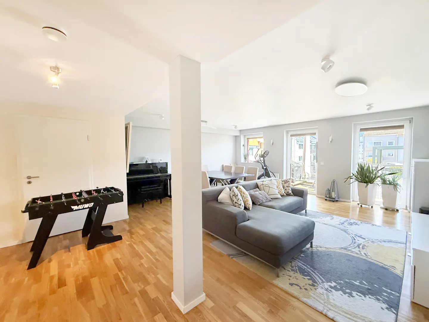 Bright, open-concept living space with a gray sectional sofa, foosball table, piano, and dining area on hardwood floors.